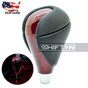 May include: A car gear shift knob with a glossy, reddish-brown wooden finish and a black leather grip. The knob has a silver base and the word "SHIFT" is visible. The image includes a smaller inset showing the knob installed.