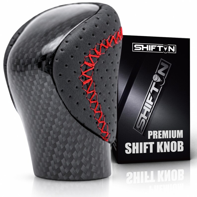 May include: A gear shift knob featuring a black carbon fibre pattern and perforated black side panels with red cross-stitching. Next to it stands a black rectangular box with the text SHIFTN and PREMIUM SHIFT KNOB printed in white.