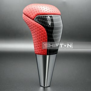May include: A red and black shift knob with a perforated leather top and carbon fiber accents. The base is a polished silver metal. The text "SHIFT N" is visible on the side.