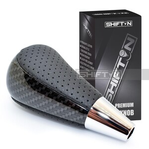 May include: A black and silver shift knob with a carbon fiber pattern and perforated leather top. The knob has a chrome base and is next to a black box with the text "SHIFT N" and "PREMIUM KNOB".