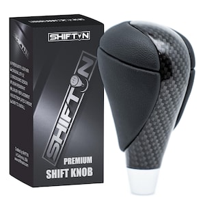May include: A black and carbon fiber shift knob with a white base, next to a black box. The box has the text "SHIFT N" and "PREMIUM SHIFT KNOB". The shift knob has a leather top and a carbon fiber body.