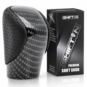 May include: A gear shift knob featuring a carbon fibre pattern and black perforated leather side panels. Next to it stands a black rectangular product box labelled SHIFTON Premium Shift Knob with a graphic of a gear pattern on the letter O.