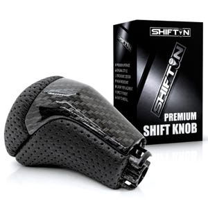 May include: A black gear shift knob featuring a carbon fiber top section and perforated black side panels. The product is displayed next to its black packaging box, which features the text SHIFTN and PREMIUM SHIFT KNOB in white lettering.