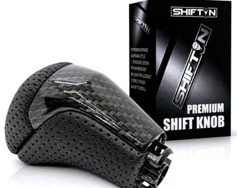 May include: A black gear shift knob featuring a carbon fibre top section and perforated black side panels. The product is displayed next to its black packaging box, which features the text SHIFTN and PREMIUM SHIFT KNOB in white lettering.