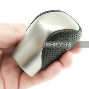 May include: A silver and black car gear shift knob. The top portion is silver, while the sides are black with small perforations. The text "SHIFT N" is visible on the side of the knob.