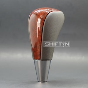 May include: A car gear shift knob with a wood grain top and a gray leather grip. The base is a polished silver color. The text "SHIFT N" is visible on the side.