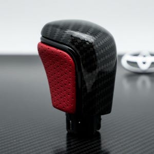 May include: A black and red car gear shift knob with a carbon fiber pattern. The red section has a textured, perforated appearance. The knob is designed for a vehicle's interior.