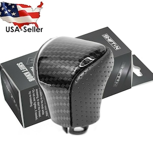 May include: A black and carbon fiber shift knob with a textured grip. The product is packaged in a black box with the text "SHIFT KNOB" and "SHIFTIN". The image also includes a USA seller graphic.