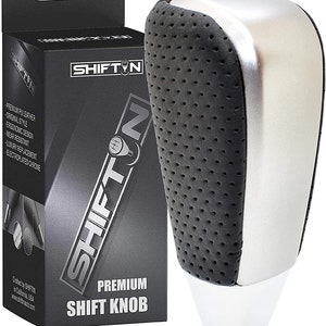May include: A silver and black shift knob with a perforated leather grip. The knob is labeled "SHIFT N" and "PREMIUM SHIFT KNOB".