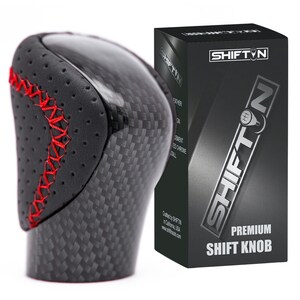 May include: A black and carbon fiber shift knob with red stitching. The shift knob has a perforated leather top and a carbon fiber weave pattern. The box next to it reads "SHIFTN" and "PREMIUM SHIFT KNOB".