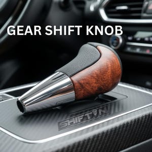 May include: A gear shift knob with a black leather top and a wood-grain handle. The base is chrome, and the knob is set in a carbon fiber console. The text "GEAR SHIFT KNOB" is displayed above the knob.