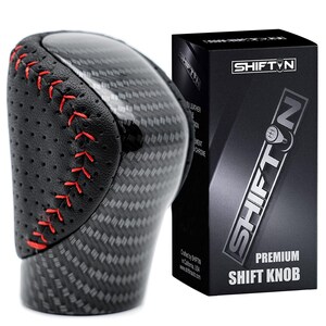 May include: A black and carbon fiber shift knob with red stitching. The shift knob has a leather top and is next to a black box with the text "SHIFTN" and "PREMIUM SHIFT KNOB".