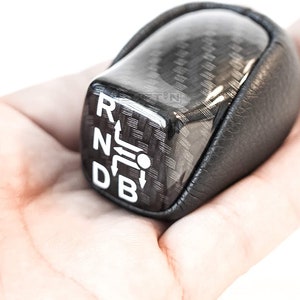 Op de afbeelding: Een zwarte en koolstofvezel versnellingspookknop met witte letters die de versnellingsposities aangeven: R, N, D en B. De knop heeft een lederen grip en wordt in een hand gehouden tegen een witte achtergrond.