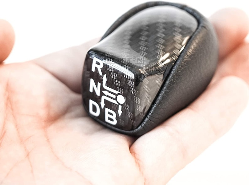 Op de afbeelding: Een zwarte en koolstofvezel versnellingspookknop met witte letters die de versnellingsposities aangeven: R, N, D en B. De knop heeft een lederen grip en wordt in een hand gehouden tegen een witte achtergrond.