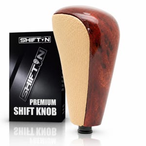 May include: A gear shift knob featuring a polished dark brown wood grain finish on the sides and a textured beige center panel. Next to it stands a black rectangular box with the text SHIFTN and PREMIUM SHIFT KNOB printed in white.