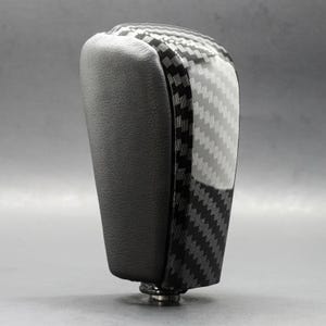 May include: A black and carbon fiber patterned car gear shift knob. The top portion has a glossy carbon fiber weave design, while the side is a matte black. The base is a silver metal.
