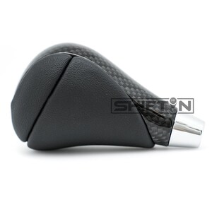 May include: A black and carbon fiber shift knob with a chrome base. The shift knob has a textured carbon fiber pattern and a smooth, black leather-like section. The word "SHIFT" is visible on the side.