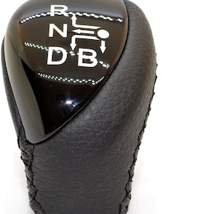 May include: Black leather gear shift knob with a black plastic top. The top has white lettering that reads "R N D B" with arrows indicating the direction to shift.