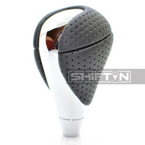 May include: A car gear shift knob with a black, perforated leather grip and a shiny chrome and white base. The word "SHIFT" is visible on the side of the knob.