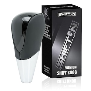 May include: A black and white shift knob with a glossy finish, next to a black box. The box has the text "SHIFTN" and "PREMIUM SHIFT KNOB". The shift knob has a black top and a white base.