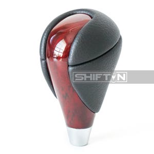 May include: A car gear shift knob with a glossy, reddish-brown wooden finish and black leather accents. The base is silver, and the text "SHIFT N" is visible on the side.