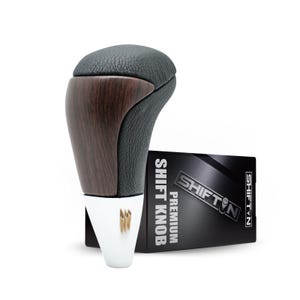 May include: A car shift knob with a black leather top and a wood grain finish. The knob has a white base with a gold emblem. A black box with the text "SHIFT KNOP PREMIUM" is next to the knob.