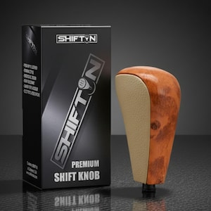 May include: A premium shift knob with a wood grain finish and tan leather side panel. The black box next to it has the text "SHIFT N" and "PREMIUM SHIFT KNOB". The shift knob is designed for a car's gear shift.