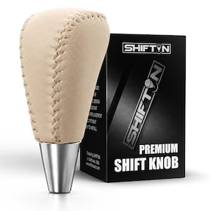 May include: A beige leather shift knob with visible stitching and a metallic silver base, positioned next to a black product box labeled SHIFTN PREMIUM SHIFT KNOB. The packaging features the brand name SHIFTN and additional text details.