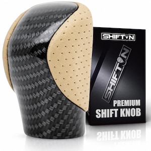 May include: A gear shift knob featuring a black carbon fibre pattern on the top and sides, accented by perforated beige side panels. Next to it stands a black rectangular box labelled SHIFTN PREMIUM SHIFT KNOB with a white logo and text.