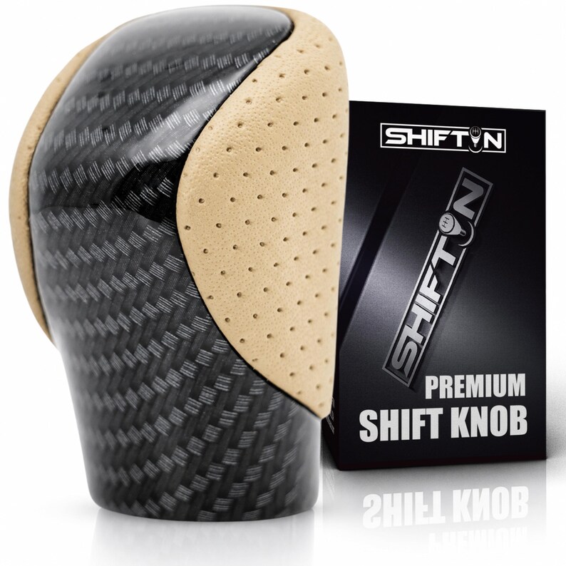 May include: A gear shift knob featuring a black carbon fibre pattern on the top and sides, accented by perforated beige side panels. Next to it stands a black rectangular box labelled SHIFTN PREMIUM SHIFT KNOB with a white logo and text.