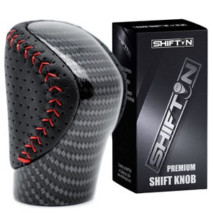 May include: A black and carbon fiber shift knob with red stitching. The shift knob is next to a black box with the text "SHIFT N PREMIUM SHIFT KNOB". The shift knob has a leather grip.