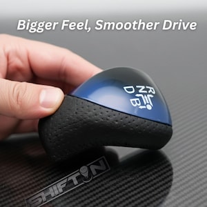 May include: A blue and black shift knob with the text "Bigger Feel, Smoother Drive". The shift knob has a glossy blue top and a textured black grip. The shift pattern is visible on the top.