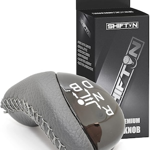 May include: A gray leather and black shift knob with white gear markings. The shift knob is next to a black box with the text "SHIFTIN" and "PREMIUM KNOB". The box also lists features like "PREMIUM PU LEATHER".