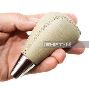 May include: A beige leather shift knob with silver metal base. The shift knob has a stitched seam along the top and the text "SHIFT N" is visible on the side.