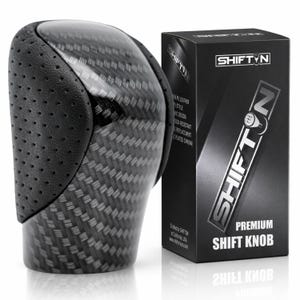 May include: A black gear shift knob featuring a carbon fiber pattern and perforated black side panels. Next to it stands a black rectangular product box labeled SHIFTON Premium Shift Knob with a graphic of a gear pattern on the letter O.
