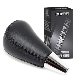 May include: Black leather shift knob with silver base. The shift knob has a stitched design and is next to a black box with the text "SHIFTN" and "PREMIUM SHIFT KNOB".