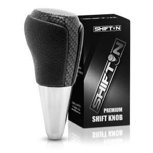 May include: A gear shift knob featuring a black textured grip, a carbon fiber pattern top, and a silver metallic base. It is displayed next to a black rectangular box labeled SHIFTN PREMIUM SHIFT KNOB with a graphic of a gear shift pattern.