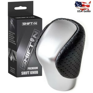 May include: A premium shift knob with a black perforated leather grip and a silver-colored base. The product is packaged in a black box with the brand name "SHIFTN" and product details. The box also has the text "PREMIUM SHIFT KNOB".