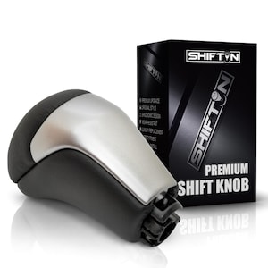 May include: A black and silver gear shift knob with a smooth, curved design. The knob is positioned in front of a black rectangular box labeled SHIFTON PREMIUM SHIFT KNOB, which features white text and a graphic of a gear shift pattern.