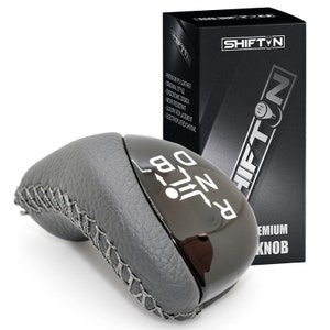 May include: Black leather gear shift knob with a chrome finish and the words "RND" and "JIL" printed on it. The knob is packaged in a black box with the word "SHIFT'N" printed on it.