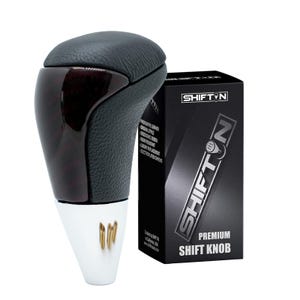 May include: A black and wood-grain shift knob with a white base and gold accents. The shift knob is next to a black box with the text "SHIFT N" and "PREMIUM SHIFT KNOB". The shift knob has a leather top.