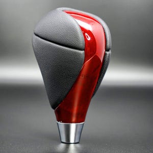May include: A car gear shift knob with a black textured leather top and a glossy red center. The base is silver-colored metal. The gear shift knob is designed for a vehicle's interior.