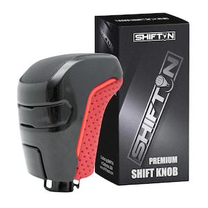 May include: Black and red shift knob with a textured grip, next to a black box. The box has the text "SHIFT N" and "PREMIUM SHIFT KNOB". The shift knob is a car accessory.