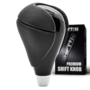 May include: A black gear shift knob with a textured leather-like upper section and a smooth, glossy black center panel. The base is a tapered silver metallic component. Beside it stands a black box labeled SHIFTON PREMIUM SHIFT KNOB.