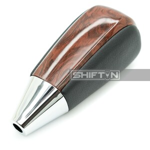 May include: A car gear shift knob with a wood grain top and a black leather grip. The bottom is chrome with the text "SHIFT N".