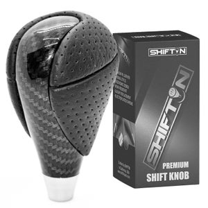 May include: A black and gray shift knob with a carbon fiber design and perforated leather grip. The product is next to a black box with the text "SHIFT" and "PREMIUM SHIFT KNOB".