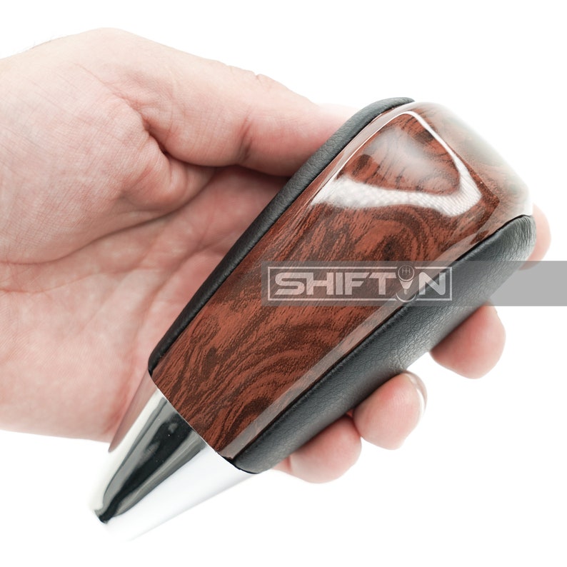 May include: A brown wood grain shift knob with a black leather wrap and a chrome tip. The knob is held in a hand.