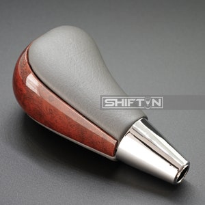 May include: A brown wood and gray leather gear shift knob with a chrome tip. The knob has a silver stripe around the middle.