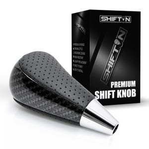 May include: A black gear shift knob featuring a perforated leather-like top section, a carbon fiber patterned side, and a polished silver-tone base. The product is displayed next to its black packaging box labeled SHIFTN PREMIUM SHIFT KNOB.