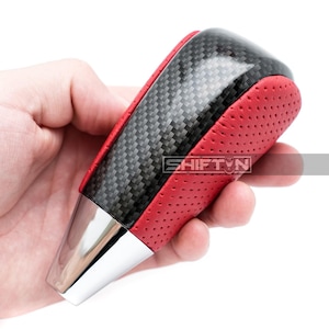 May include: A car gear shift knob with a carbon fiber top and red perforated sides. The base is silver, and the text "SHIFT N" is visible on the side.